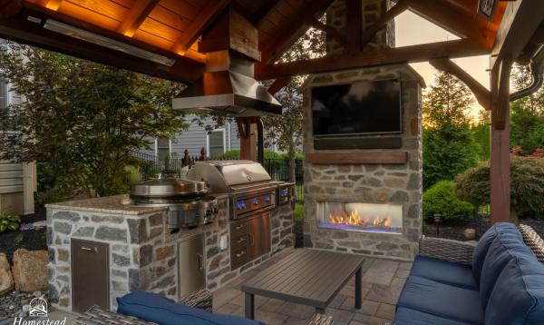 A cozy outdoor kitchen with a stone grill, stainless steel appliances, a flat-screen TV mounted above a fireplace, and a seating area with a blue cushioned wicker sofa and coffee table. The setup is under a 12' x 14' timber frame pavilion with warm lighting and surrounded by greenery.