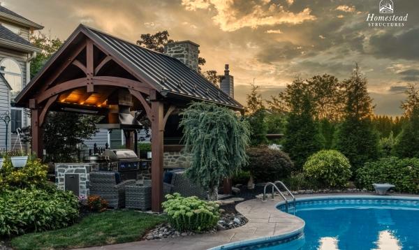 A backyard space features a pool with curved edges, surrounded by lush greenery and landscaping. Nearby, a 12' x 14' timber frame pavilion with a stone base and seating area stands under a cloudy sunset sky.