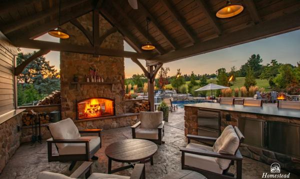 A 16' x 20' timber frame pavilion featuring cushioned seating around a round coffee table, a stone fireplace with a lit fire, and a view of a lush garden with a pool.