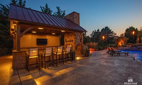 A 16' x 20' timber frame pavilion with a built-in stone fireplace, and the area is softly lit with warm lights. Nearby, lounge chairs are placed beside a glowing pool. Trees and torches add to the serene ambiance.