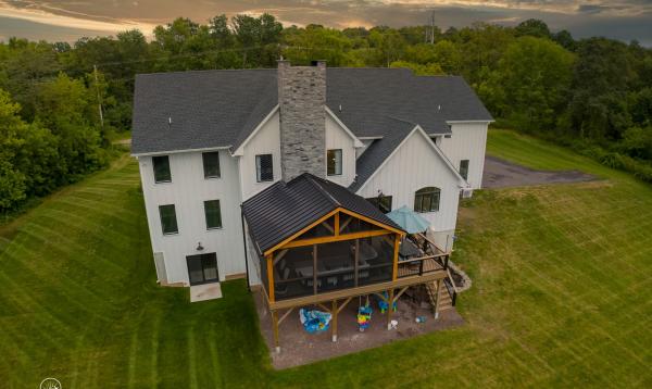 A large two-story house with a white exterior and black roof is surrounded by a well-manicured lawn and trees.  A 20' x 20' Attached timber frame pavilion with 580sq ft TImbertech Deck & Stairs are visible at the rear of the house.