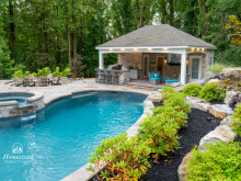 Elegant stone pool pavilion with spacious entertainment area including an outdoor kitchen, built-in grill, comfortable seating, fire pit area, and integrated landscaping, perfect for gatherings